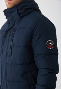 Námořnická puffer bunda má vysoký límec, zapínání na zip na předních kapsách a kulatou nášivku na levém rukávu. Hladká, izolační tkanina.