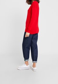 Pull en tricot rouge, coupe décontractée, décolleté rond. Associé à un jean en denim bleu foncé, avec ourlets, et des baskets blanches avec des accents noirs.