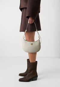 Structured, light gray leather handbag with a curved shape, gold hardware, and an adjustable strap, held by a person in a brown outfit and boots.