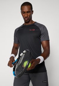 T-shirt de sport noir à manches courtes avec des accents gris, arborant un logo rouge. La personne tient une raquette de tennis et une balle de tennis jaune.