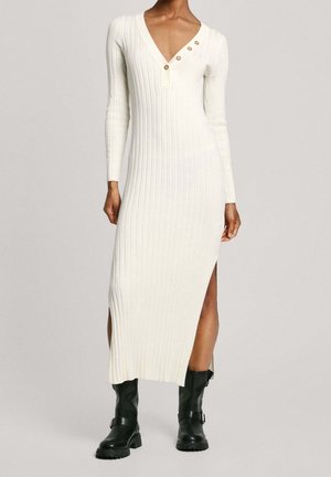 Długa biała sukienka z prążkowanego dzianiny, z dekoltem w serek i guzikami, noszona z czarnymi skórzanymi kozakami sięgającymi do połowy łydki, stojąca na jednolitym tle.