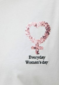 Witte katoenen shirt met een roze pailletten hart en vrouwelijk symbool. Zwarte geborduurde tekst leest: "Elke dag is Vrouwendag."