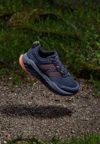 Grijze sport schoen met oranje accenten, bovenmateriaal van mesh en gevoerde zool, boven een grindachtige ondergrond zwevend met groene achtergrond.