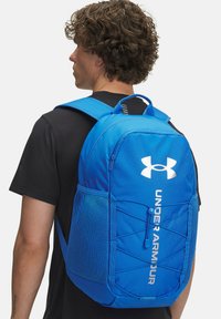 Modrá batoh Under Armour s zaobleným tvarom, sieťovými bočnými vreckami, elastickými šnúrkami a bielou potlačou loga na prednej strane. Nastaviteľné popruhy.