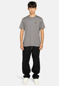 Grijze katoenen t-shirt met een ronde hals en een klein embleem op de linkerborst, gecombineerd met loszittende zwarte jeans en witte sneakers.