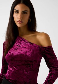 Schulterfreies Top aus tiefmagentafarbenem Samt mit langen Ärmeln und einem weichen, strukturierten Finish. Die glatte Silhouette betont den Ausschnitt.