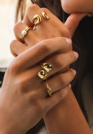 Manos entrelazadas que llevan tres anillos de oro, uno con una perla blanca y dos con pequeñas cuentas plateadas, sobre piel de tono marrón claro.