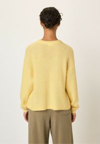 Rumena puhasta majica z ohlapnim krojem, okroglo ovratnikom in dolgimi rokavi, ki jo dopolnjujejo rebrasti manšeti in spodnji rob. V kombinaciji z teksturiranimi bež hlačami.