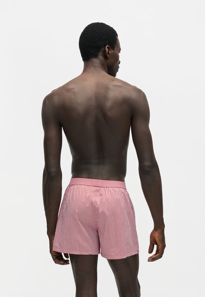 Άνδρας με σκούρο δέρμα που φαίνεται από πίσω, φοράει κόκκινα και λευκά κατακόρυφα ριγωτά σορτς, στέκεται μπροστά σε μια απλή ανοιχτή επιφάνεια.