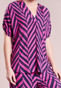 Blusa a righe chevron rosa e blu navy con maniche corte, scollatura a V e vestibilità comoda. Tessuto leggero con una texture morbida.