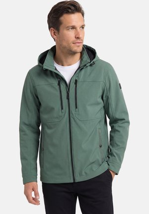 Man wearing green hooded jacket with black zippers over white shirt, looking right, hands relaxed by sides.