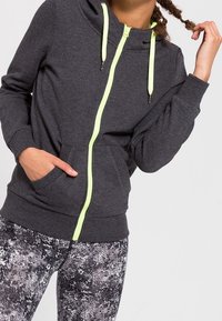 Sudadera gris con cremallera y cordones en verde neón, con un bolsillo delantero, combinada con leggings de patrón en tonos de negro y gris.
