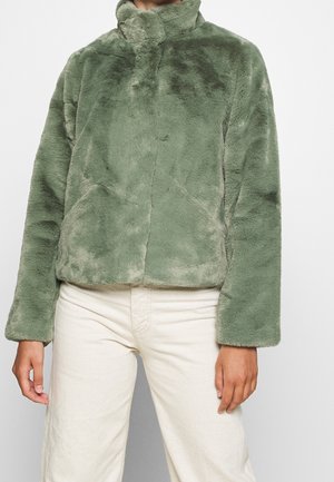 Personne portant une veste douce et pelucheuse vert olive clair avec des manches longues et un col montant, associée à un pantalon écru.