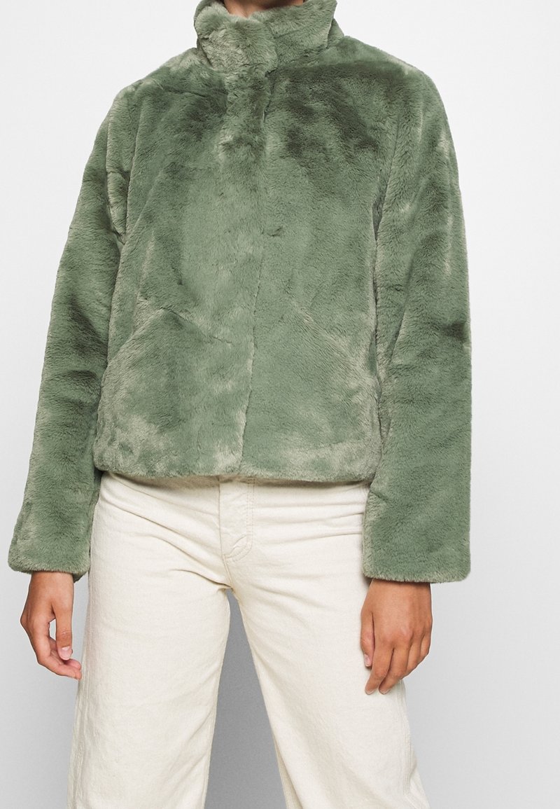 Personne portant une veste douce et pelucheuse vert olive clair avec des manches longues et un col montant, associée à un pantalon écru.