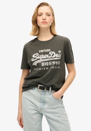 T-shirt en coton vert foncé avec le texte imprimé en blanc "SuperDry" et des accents. Assorti à un jean bleu clair et une ceinture noire avec une boucle en argent.
