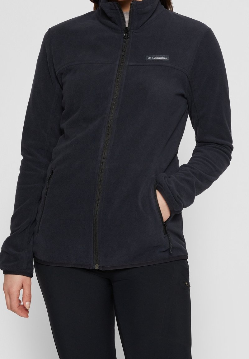 Tummansininen fleecetakki, jossa on vetoketjullinen etuosa, korkea kaulus, sivutaskut ja pehmeä tekstuuri. Rinnassa on logo.