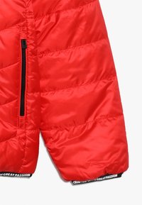 Manga de una chaqueta acolchada roja con una cremallera negra y puño elástico que presenta el texto blanco "ELABORADO CON GRAN PASIÓN."