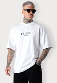 T-shirt en coton blanc à manches courtes avec un logo imprimé "ESTEEM MARBELLA". Porté avec un pantalon noir et des lunettes de soleil foncées.