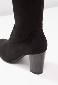 Bottine en daim noir avec un bout arrondi et un talon haut texturé. Le design présente des lignes épurées et une silhouette ajustée.