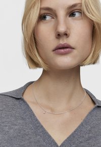 Collana d'argento con una catena delicata e un piccolo pendente, indossata sopra una maglietta grigia lavorata a maglia con scollatura a V. Presenta una texture liscia.