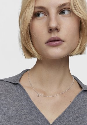 Collar de plata con una cadena delicada y un pequeño colgante, llevado sobre una camiseta de punto gris con escote en V. Presenta una textura suave.