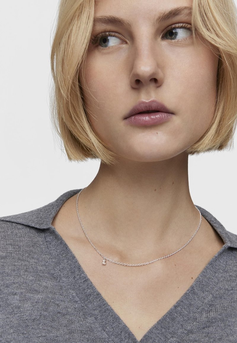 Collana d'argento con una catena delicata e un piccolo pendente, indossata sopra una maglietta grigia lavorata a maglia con scollatura a V. Presenta una texture liscia.