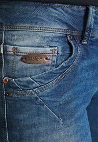 Un primer plano de unos vaqueros de mezclilla azul que presentan una placa de logo de metal, remaches de cobre y costuras en contraste, mostrando una textura y un diseño deslavado.