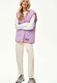 Kobieta z długimi blond włosami, ubrana w fioletową pikowaną kamizelkę z pomarańczową lamówką, białą koszulę z długim rękawem, jasnobeżowe spodnie i trampki.