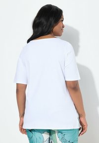 Camiseta blanca de manga corta con escote barco, corte relajado y aberturas laterales. La tela parece suave, complementando unos pantalones de teal con estampado.