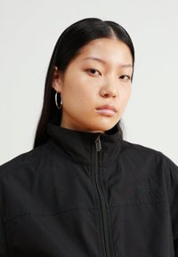 Svart zip-up jakke med høy krage, med logo på venstre side og glatt tekstur, båret av en person med glatt, mørkt hår.