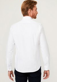 Camicia bianca a maniche lunghe con colletto classico, dotata di polsini a bottoni, una texture liscia e un orlo curvo, indossata con jeans scuri.