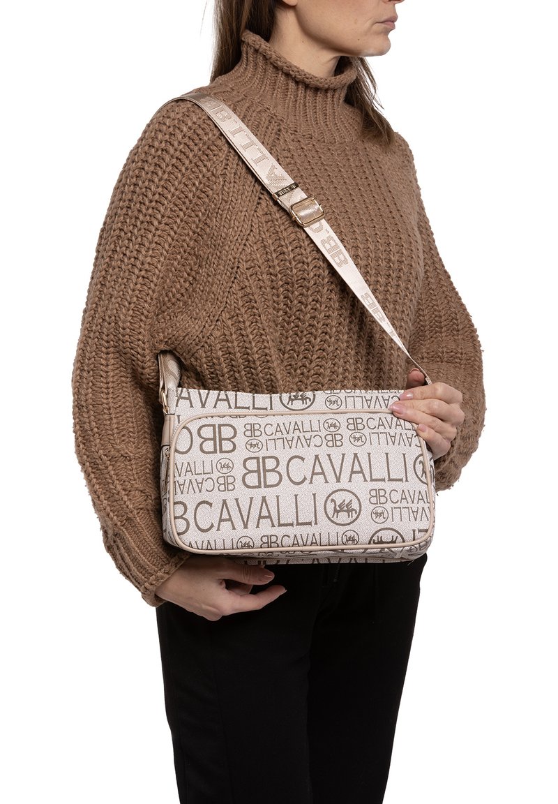 Borsa a spalla beige con logo, realizzata in tessuto texturizzato, con dettagli dorati, design sottile e tracolla regolabile, indossata sopra un maglione marrone lavorato a maglia.