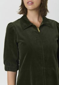 Mujer vistiendo un vestido de terciopelo verde oscuro con mangas abullonadas hasta el codo y una cremallera dorada en el cuello, mostrado de los hombros a la cintura.