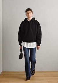Svart oversized hettegenser lagd over en hvit skjorte, kombinert med vide blå jeans og mønstrede sko, bærende en svart håndveske.