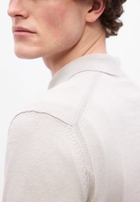 Κοντινό πλάνο ενός ατόμου που φοράει ανοιχτό μπεζ πλεκτό πουλόβερ με λεπτομερή ραφή ραφής κατά μήκος του ώμου και της πλάτης.