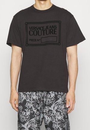 Uomo che indossa una t-shirt nera Versace Jeans Couture e pantaloncini bianchi e neri con scritte e motivi floreali.