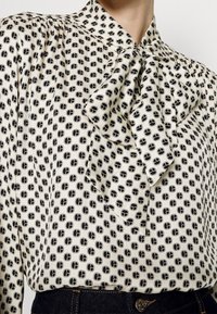 Beige bluse med sort geometrisk mønster, der har en knyttet hals-bue, båret ind i højtaljede mørk denim jeans.