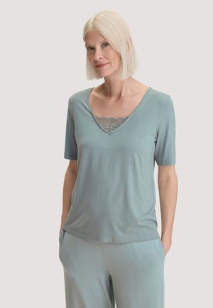 Frau mit hellblonden Haaren trägt ein hellblaues V-Ausschnitt-Shirt mit Spitzen-Detail und passende Hose, steht mit den Händen in den Taschen.