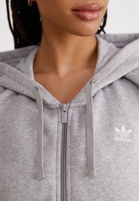 Šedá mikina na zip vyrobená z měkkého materiálu, s kapucí na šňůrku, jemným logem a hladkým zipem se stříbrným táhlem.