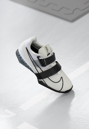 Chaussure de sport blanche et noire avec deux larges bandes noires et une semelle visible amortie par air sur un sol gris clair.