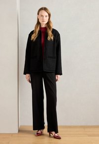 Veste noire texturée à l'avant ouvert et avec deux poches, portée sur un haut bordeaux, associée à un pantalon oversized noir et des chaussures à talons rouges.