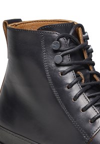 Stivale alla caviglia in pelle nera con una texture liscia, interno marrone a contrasto, lacci tondi e due bottoni decorativi sul lato esterno.