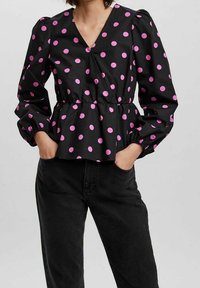 Blusa nera con pois rosa, scollo a V profondo, vita elastica e maniche a sbuffo. Esibita con jeans neri per contrasto.