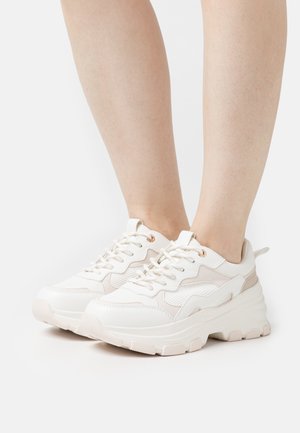 Personne portant des baskets épaisses de couleur écrue avec des semelles épaisses et une fermeture à lacets, debout devant un fond blanc uni.