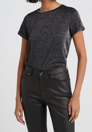 Frau trägt ein kurzärmeliges, schwarz-grau gesprenkeltes T-Shirt, das in glänzende, hoch taillierte schwarze Hosen mit Vorder Taschen gesteckt ist.