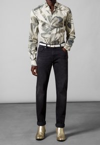 Camisa con estampado floral en tonos crema y gris, combinada con jeans negros y botines metálicos dorados, con un cinturón blanco.