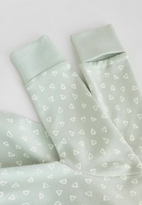 Ljust grönt tyg med ett upprepande triangulärt mönster i krämfärg. Manschetterna är i hel ljust grönt, mjukt textil med en åtsittande design.