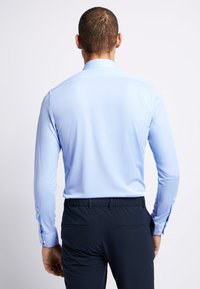Lichtblauwe longsleeve shirt met een kraag, gladde textuur en knoopdetails op de manchetten, gecombineerd met donkere broek, gezien vanuit de achterkant.