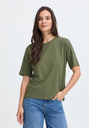 Jeune femme aux longs cheveux foncés portant une chemise à manches courtes vert olive et un jean bleu, debout avec une main dans la poche.