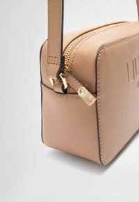 Sac à main rectangulaire beige en matériau synthétique texturé, avec une fermeture éclair dorée et une bandoulière détachable. Présente une marque en relief sur le devant.